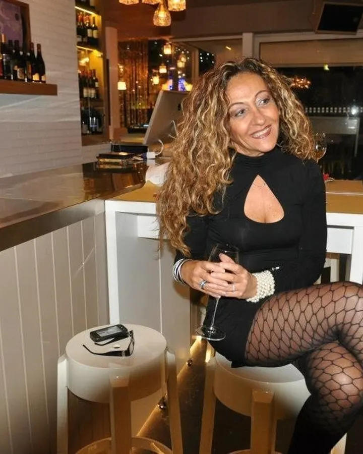 Femme aux cheveux blonds, souriante, portant une robe noire, debout dans une chambre avec un grand meuble en bois et un miroir.