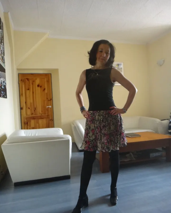 Femme aux cheveux bruns-noirs, souriante, portant une robe noire, assise sur un canapé beige, dans une pièce avec des rideaux blancs.