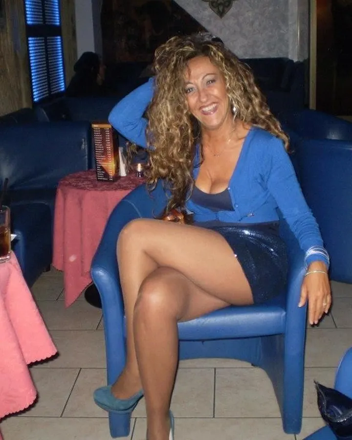 Femme blonde en robe grise, assise sur un lit avec un verre à la main