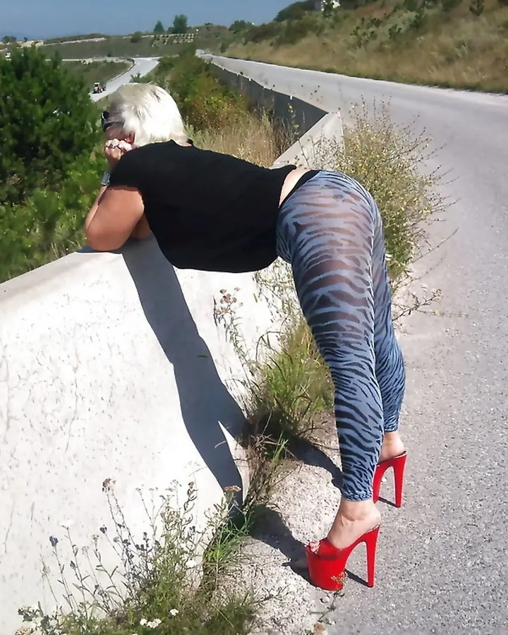 Femme aux cheveux platine courts, vêtue de noir, en position accroupie sur un bord de route, talons rouges, regard vers l'arrière.