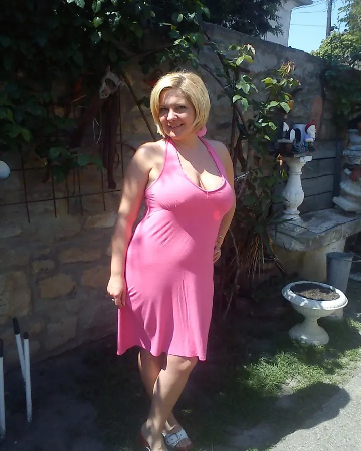 Femme aux cheveux blonds, robe rose, debout dans un jardin, sourire chaleureux, soleil en arrière-plan.