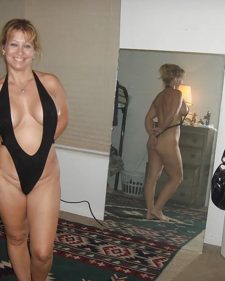 Femme aux cheveux blonds, portant un haut rouge, debout devant un miroir, les mains sur les hanches, souriant à l'appareil
