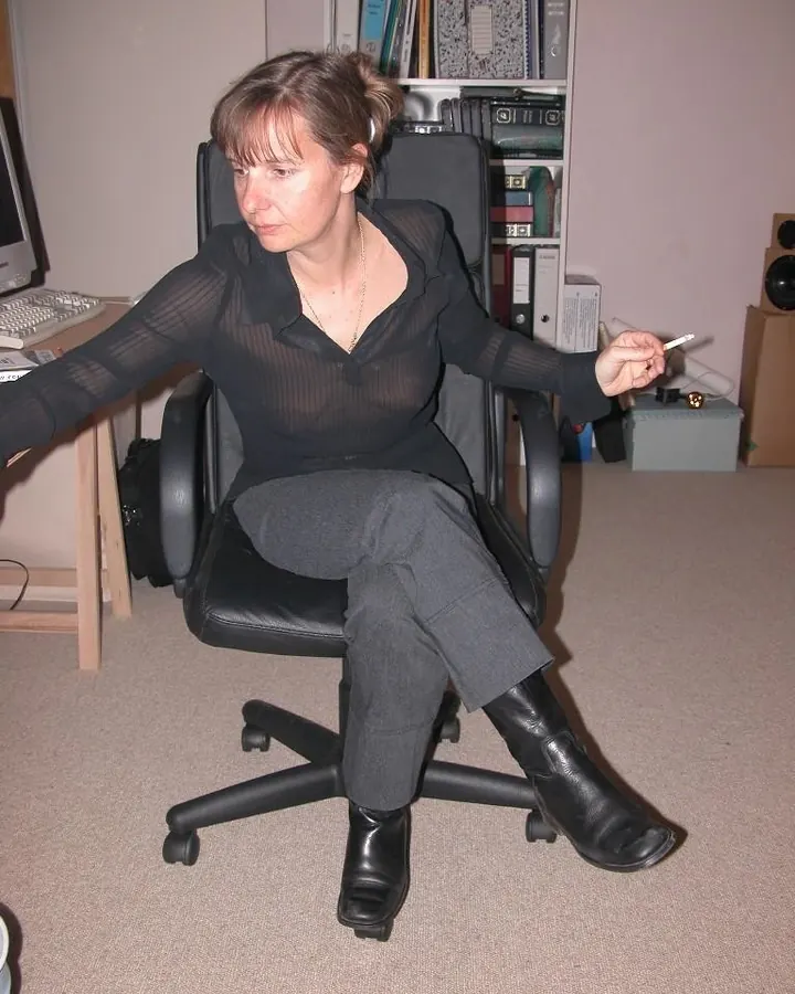 Femme aux cheveux châtains, portant une chemise noire transparente et un pantalon gris, assise sur une chaise de bureau, tenant une cigarette