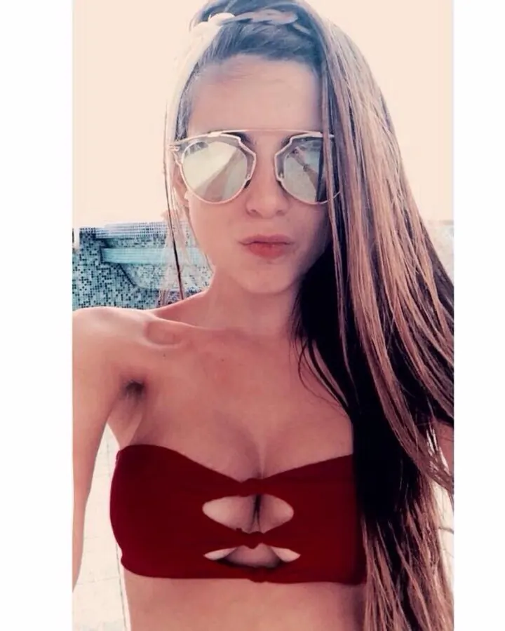 Femme aux cheveux blonds, portant un haut rouge sans manches, debout sur une plage rocheuse, regardant vers l'appareil
