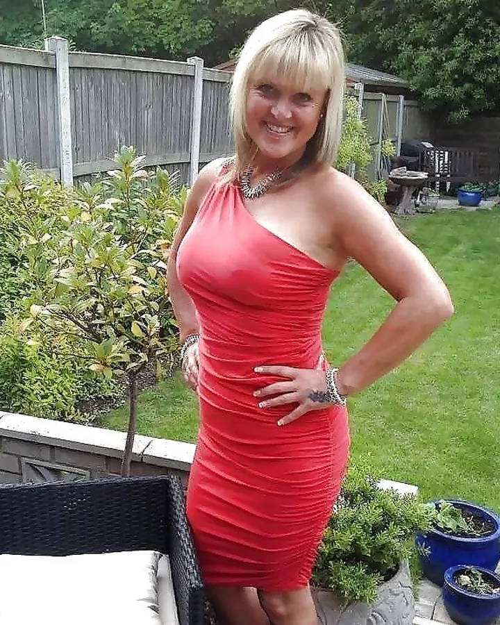 Femme blonde aux cheveux courts, portant une robe rouge ajustée, debout dans un jardin verdoyant, souriant à l'appareil