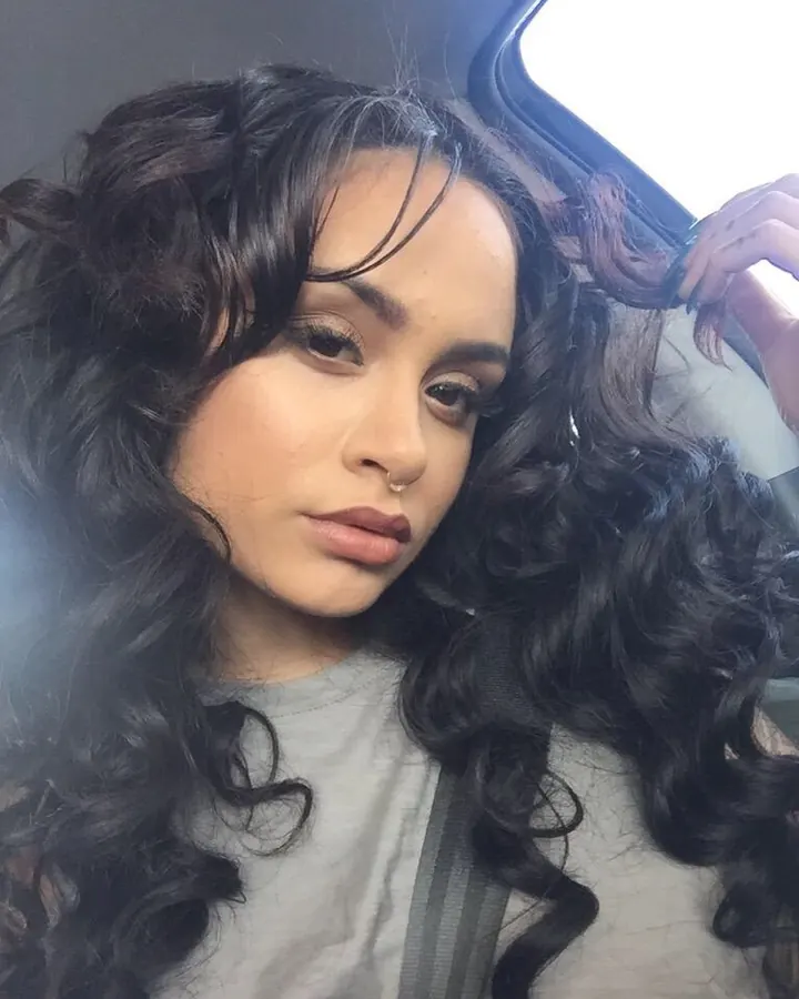 Femme aux cheveux noirs bouclés, portant un t-shirt gris, assise dans une voiture, regardant vers l'appareil