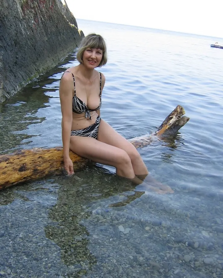 Femme blonde en maillot rayé assise sur un tronc dans l'eau claire près d'une roche