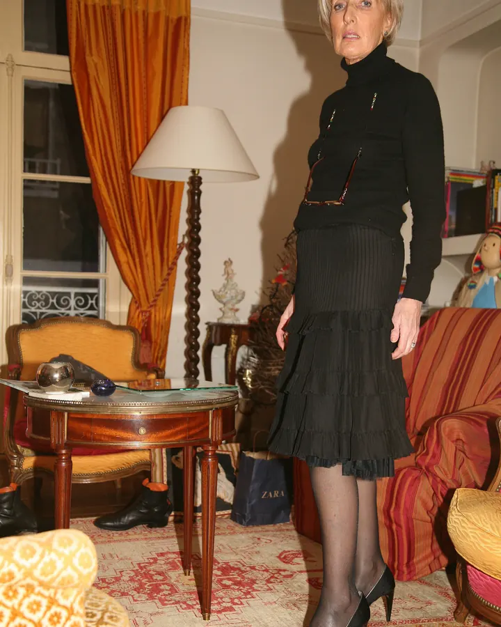 Femme aux cheveux gris courts, portant une robe noire à pois, assise dans un fauteuil beige, dans un salon avec lampe et tapis coloré.