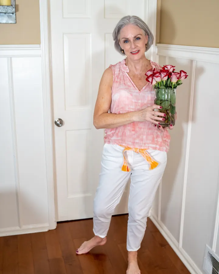 Femme aux cheveux blancs courts, portant un haut noir à col roulé, jeans bleus, chaussures beige, debout devant un bureau avec ordinateur, dans un salon élégant avec plantes et tableau.
