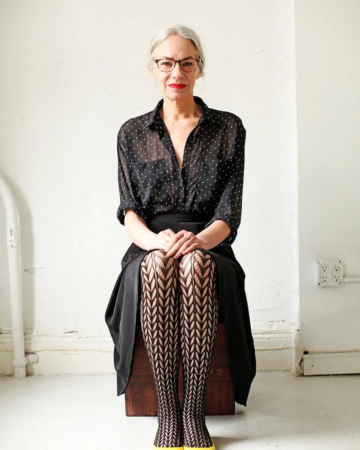 Femme aux cheveux blancs, lunettes, chemise noire à pois transparente, collants à motif géométrique et chaussures jaunes, assise sur un banc en bois dans un espace blanc.