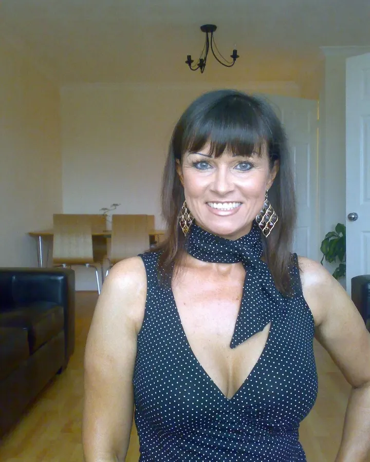 Femme aux cheveux bruns foncés, souriante, portant une robe noire à pois blancs avec un col noué, dans un salon lumineux avec canapé noir et table en bois.