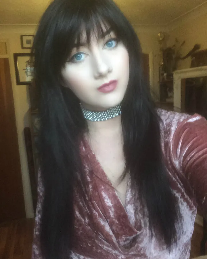 Femme brune en robe noire, posant dans un salon avec un canapé beige.