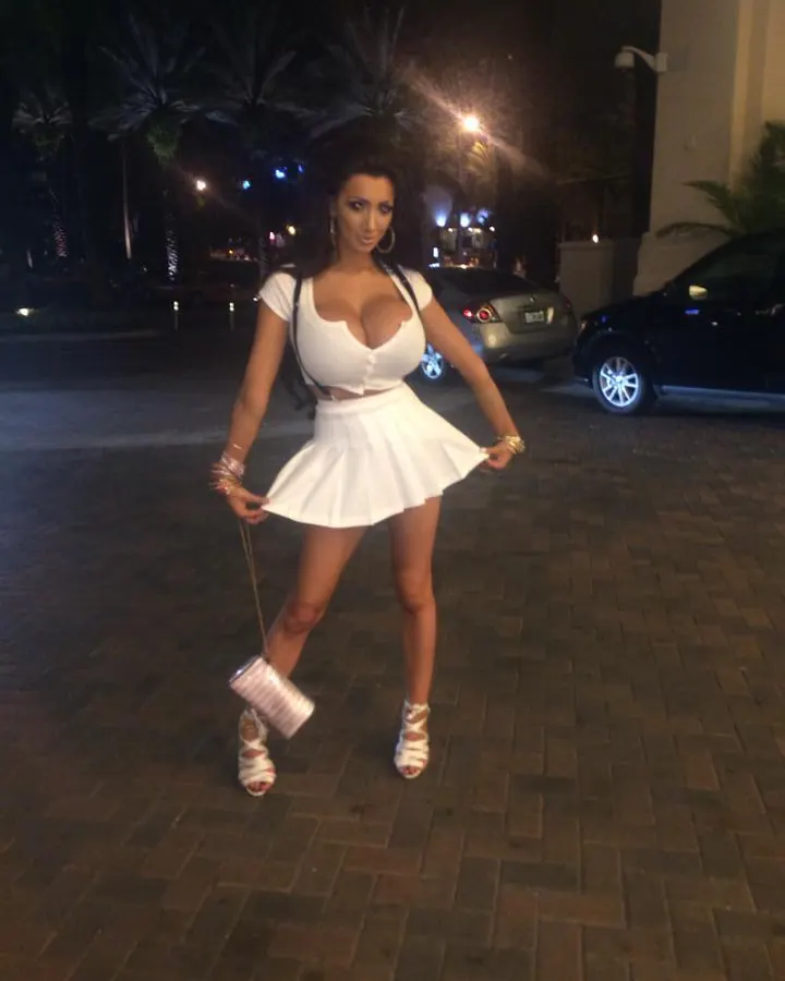 Femme aux cheveux bruns portant une tenue blanche et une jupe courte, debout la nuit près d'une voiture