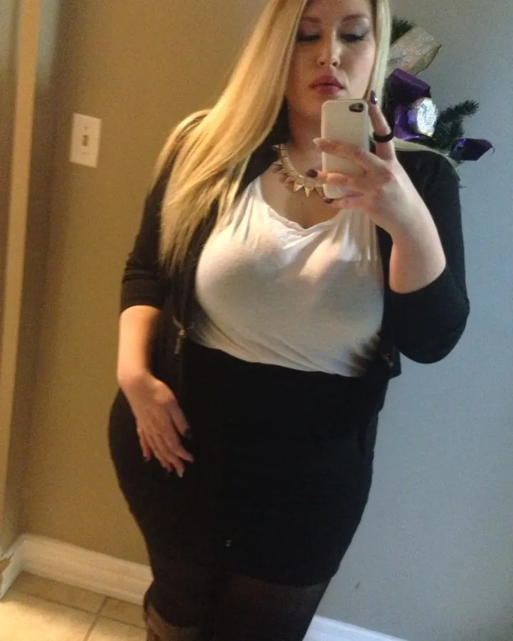 Femme blonde en tenue noire et blanche prenant un selfie dans un miroir, regard baissé.