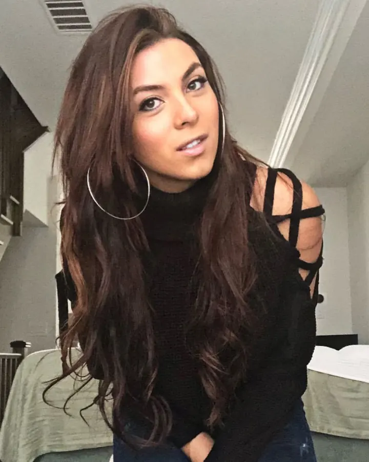 Femme brune aux cheveux longs, portant un haut noir à col en dentelle, debout dans une pièce avec des meubles en bois clair.