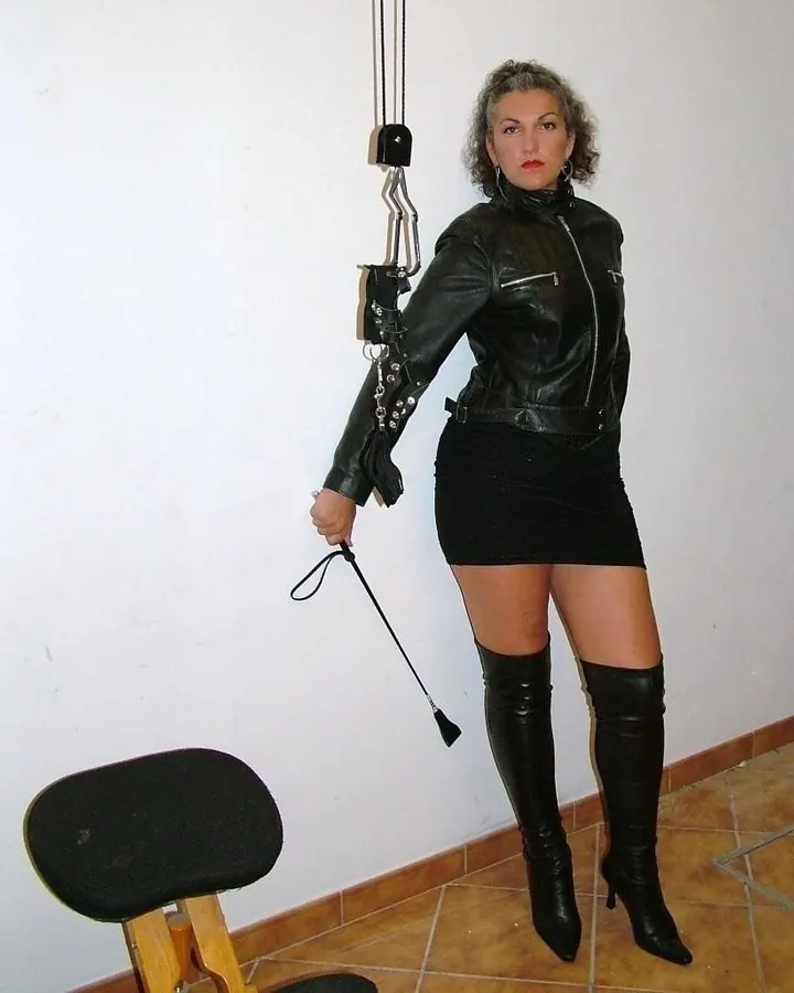 Femme blonde aux cheveux longs, portant un haut noir à col en dentelle, debout dans une pièce avec des meubles en bois clair.