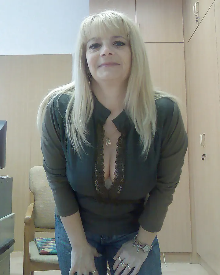 Femme blonde aux cheveux longs, portant un pull gris et un short déchiré, assise contre un mur de briques avec des tatouages visibles sur ses jambes.