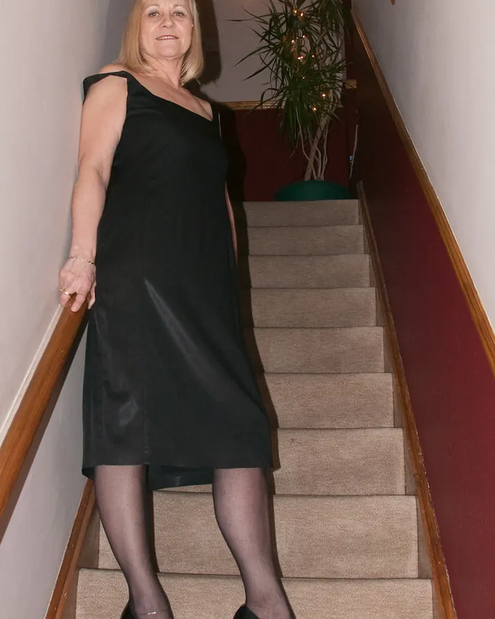 Femme blonde en robe noire, descendant un escalier, souriante, pieds en talons noirs.
