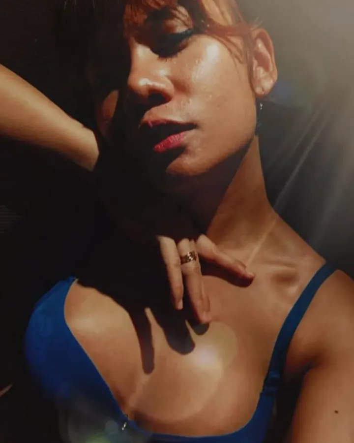 Femme aux cheveux châtains en soutien-gorge bleu, main posée sur le cou, lumière chaude et contrastée.