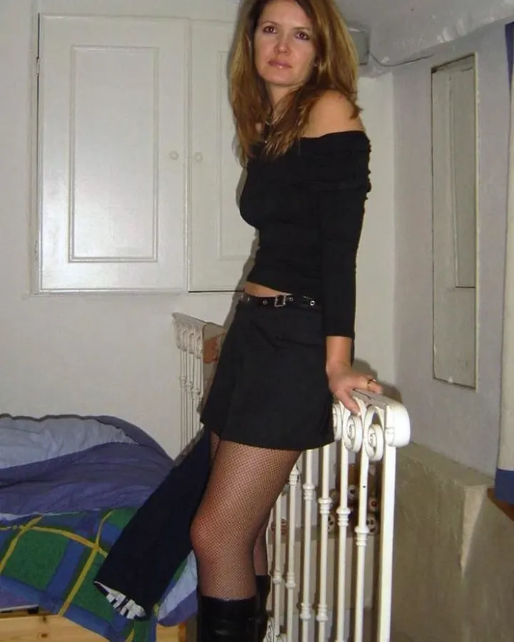 Femme en robe noire à épaules dénudées, tenant une rampe blanche dans une chambre.