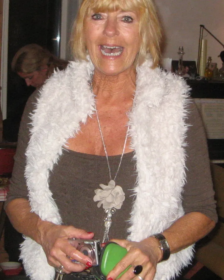 Femme en pull blanc à col roulé, souriante, tenant un appareil photo numérique et un objet vert.