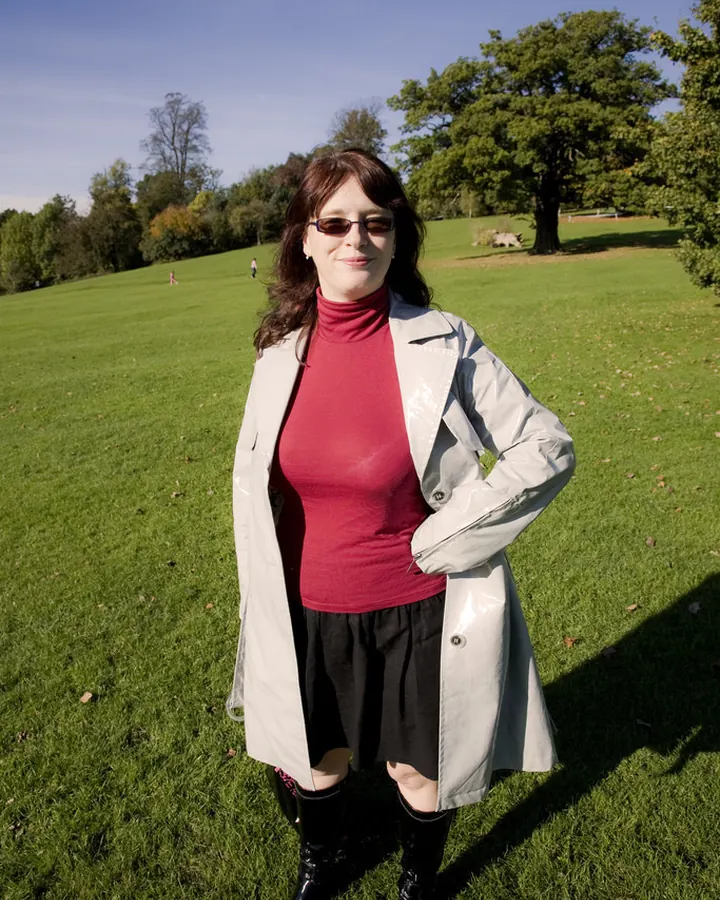 Femme en veste claire et haut rouge sur une pelouse, souriante sous un ciel bleu.
