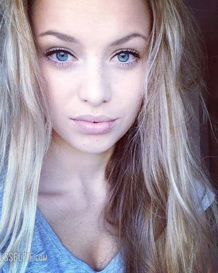 Jeune femme aux cheveux blonds platine et yeux bleus, en gros plan, regard direct, vêtue d'un haut bleu clair.