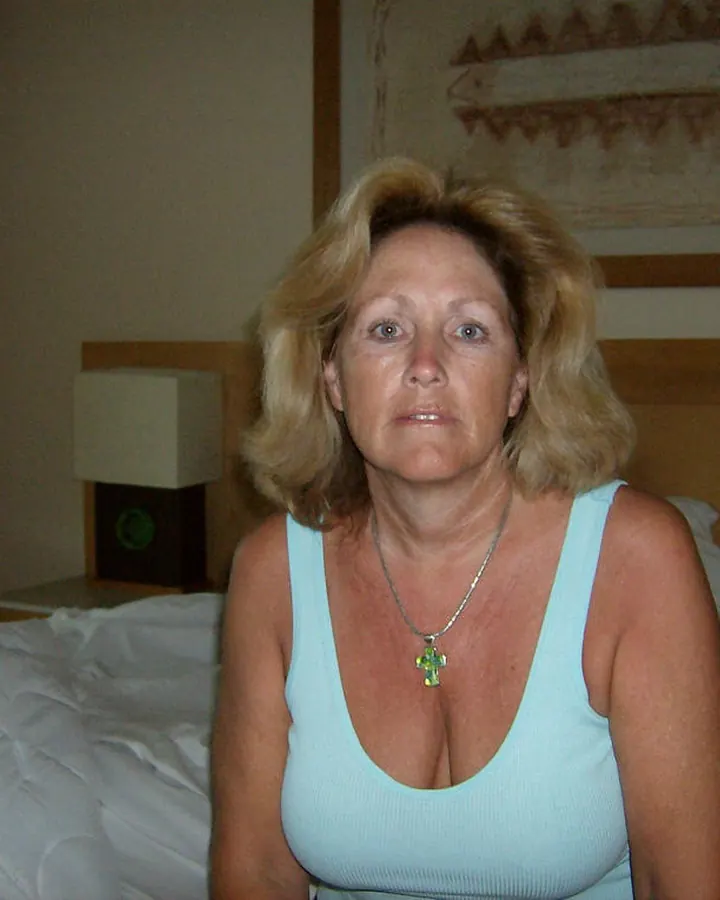Femme aux cheveux blonds courts, vêtue d’un haut blanc à volants, prenant un selfie devant un miroir, souriant légèrement.
