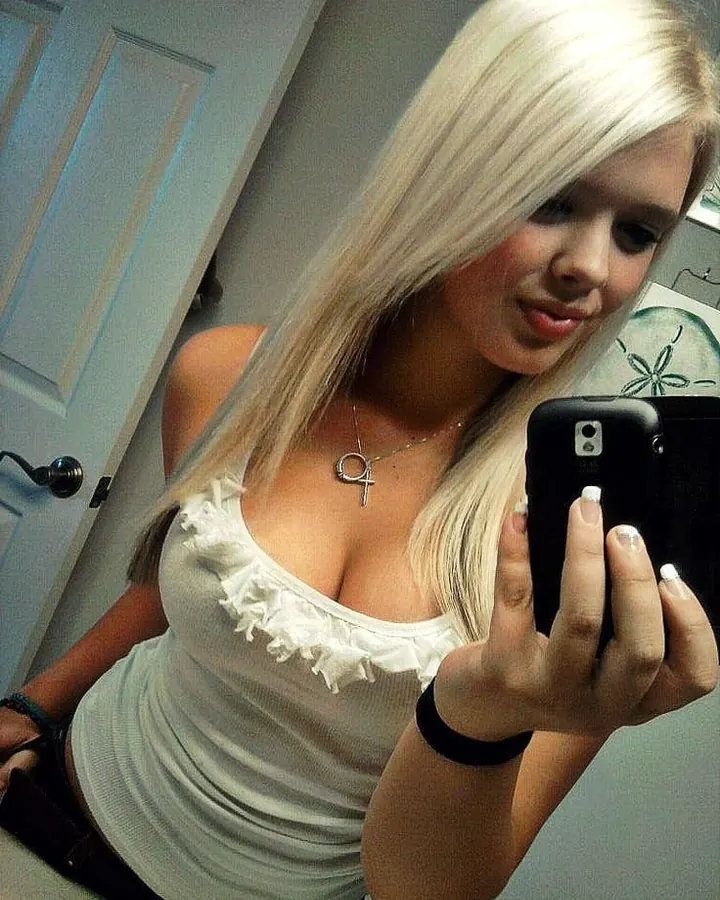 Femme blonde, vêtue d’un haut noir, prenant un selfie dans un intérieur moderne, regardant l'appareil avec un sourire discret.
