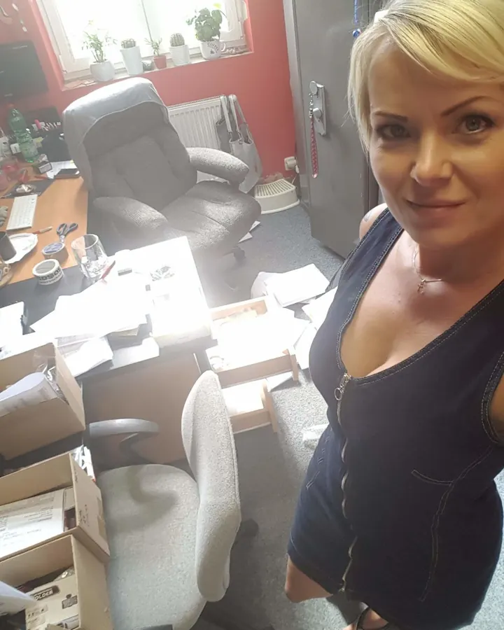 Femme blonde platine, yeux bleus, en robe noire transparente, collants noirs, escarpins noirs, assise sur une chaise dans une chambre avec bibliothèque et ordinateur.