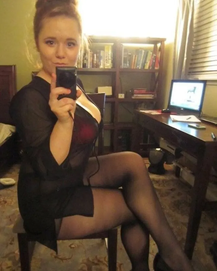 Femme blonde, yeux bleus, en robe noire transparente, collants noirs, escarpins noirs, assise sur une chaise dans une chambre avec bibliothèque et ordinateur.
