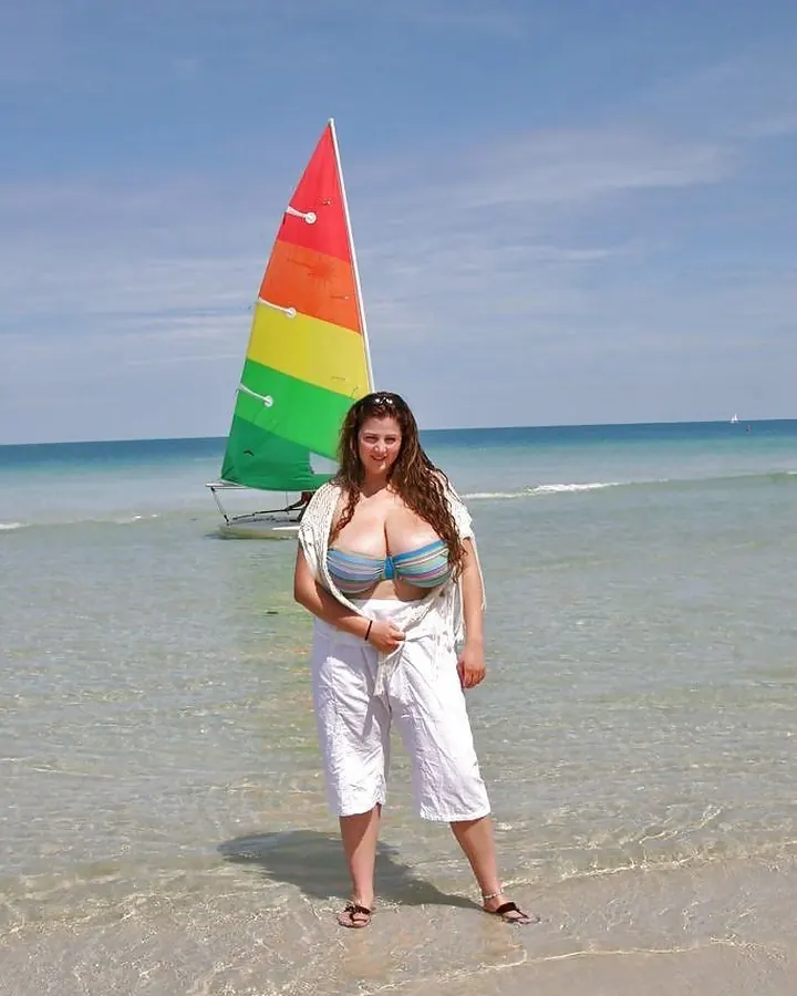 Femme en bikini multicolore, vêtue d’un pantalon blanc, debout dans l’eau peu profonde d’une plage, avec un voilier coloré en arrière-plan.