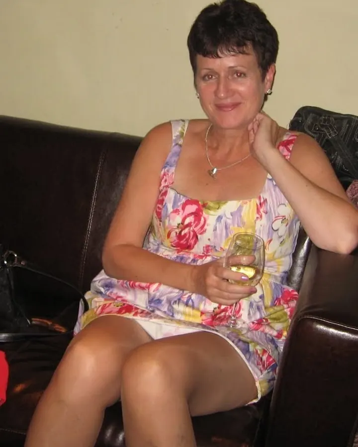 Femme aux cheveux courts, en robe fleurie, assise sur un canapé en cuir, tenant un verre de vin.