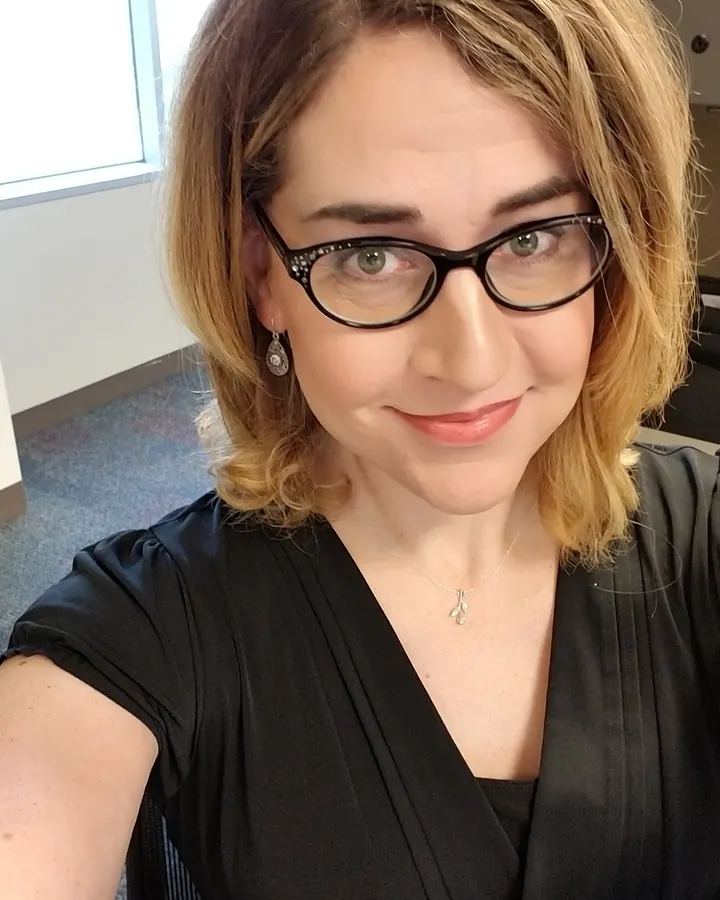 Femme aux cheveux blonds courts, portant des lunettes noires et une robe noire, souriant légèrement devant une fenêtre.