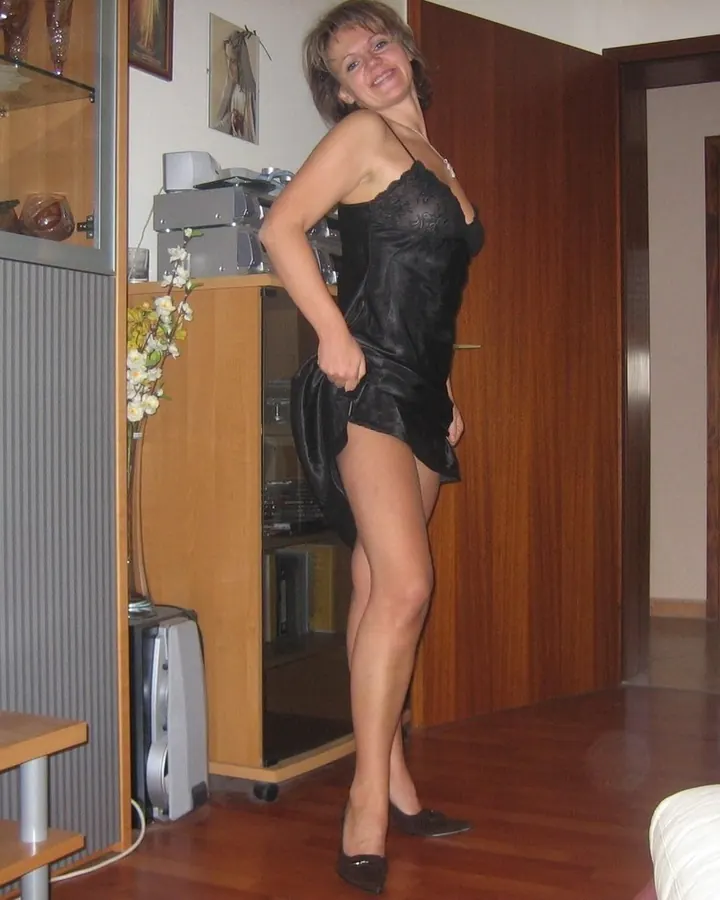 Femme blonde en robe noire courte, posant dans un salon moderne avec un téléviseur et des meubles en bois.