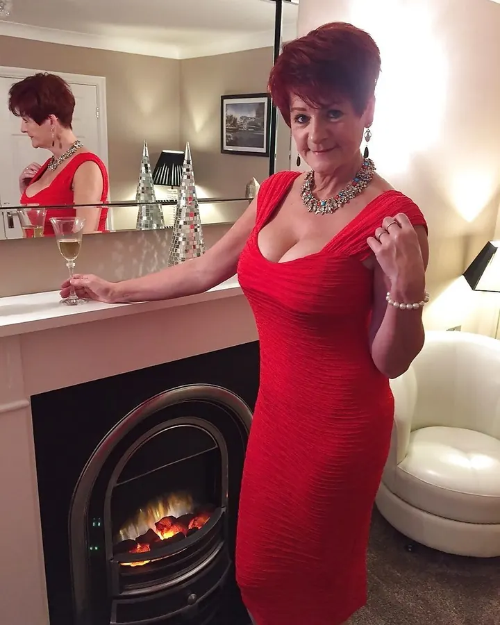 Femme aux cheveux roux courts, portant une robe rouge ajustée, tenant un verre de vin blanc près d'une cheminée allumée.