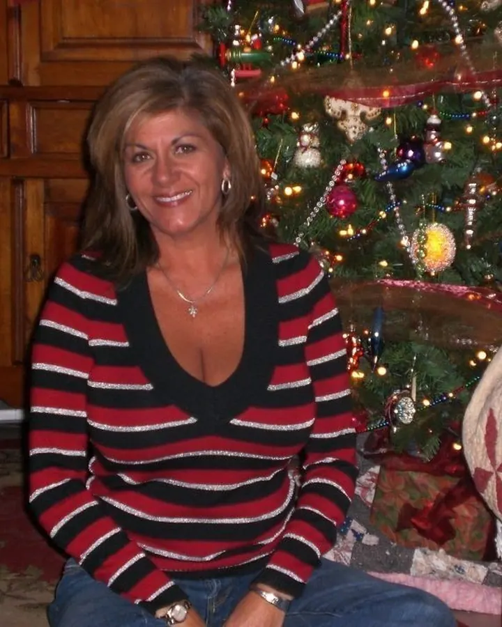 Femme aux cheveux châtains, portant un pull rayé rouge et noir, assise devant un sapin de Noël illuminé.