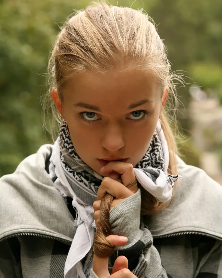 Jeune femme aux cheveux blonds, portant un sweat gris et un foulard noir et blanc, tenant une mèche de cheveux entre ses doigts, regardant fixement l'objectif.