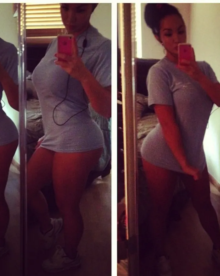 Femme africaine aux cheveux bruns, portant un haut gris et un short, prenant un selfie devant un miroir dans une chambre.