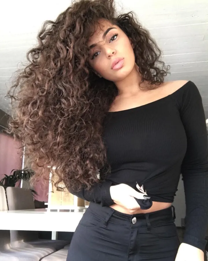 Femme latino aux cheveux bruns, portant un haut noir et un short, posant sur une plage avec un regard direct.