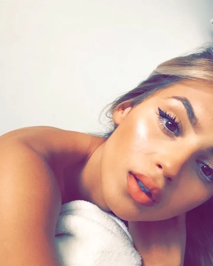 Femme aux cheveux blonds et yeux sombres, allongée, tenant une serviette blanche près de son cou, dans un cadre intime et doux.